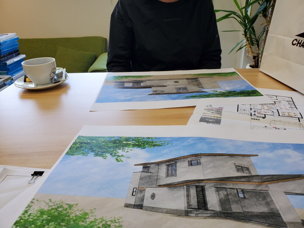 設計士との住宅プラン打ち合わせ風景。ガレージ付きオシャレモダンな二階建て住宅の完成予想パースと詳細な平面図を机に広げながら、新築計画について検討中。奈良県での注文住宅設計・建築相談のリアルな一場面。住まいのビジョンを丁寧に描くプロセスが感じられる。