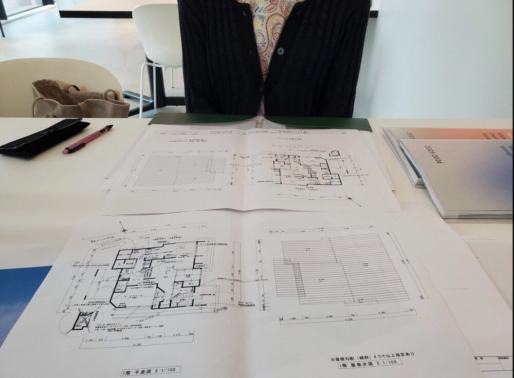 住宅の新築計画打ち合わせ風景。白いテーブルに広げられた建築設計図（平面図・屋根伏図）を囲みながら、建築家と住まい手が間取りや生活動線について検討を進めている。住まいは自然と調和する平屋を計画中で、外構や庭との関係性も含めた設計が話し合われている。設計図面には手書きの補足やスケッチも添えられ、暮らしのイメージが共有されている様子がうかがえる。奈良県の風土に根ざした和モダン住宅の設計プロセス。