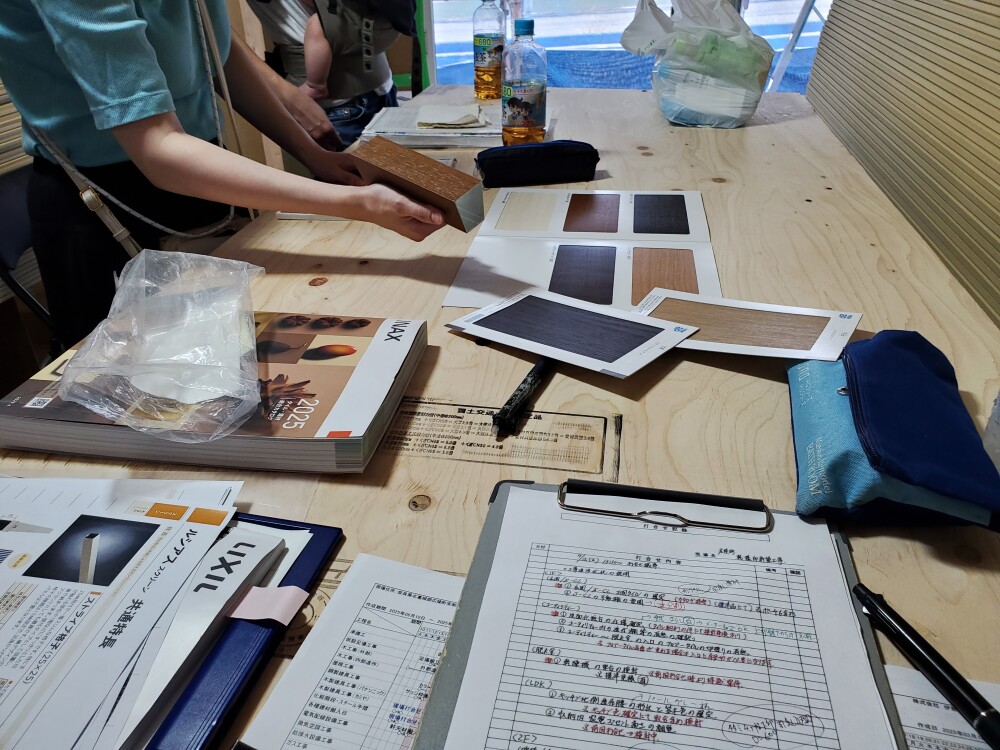 現場打合せの様子。木製サンプルやタイル、壁紙サンプルが机上に並び、建築家と住まい手が自然光の中で仕上材の選定を行っている。実物の質感と光の反射を見極めながら最終決定を進める重要な工程。