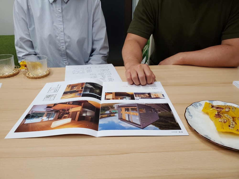 やまぐち建築設計室の打ち合わせ風景。新築住宅を検討中のご夫婦が建築家と資料を見ながら、和モダンでおしゃれな暮らしを叶える住まい計画について相談している様子。図面や施工事例の写真集を広げ、夫婦二人にちょうどいい間取りやリビングの自由な使い方、DJブースを備えた空間設計を検討している場面。