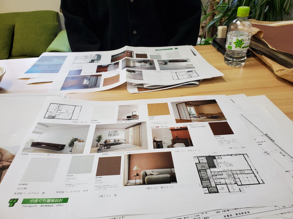 やまぐち建築設計室の打ち合わせ風景。新築住宅の間取り図やインテリアコーディネート資料を机に広げ、壁紙やクロス、家具の色合いを比較検討している様子。壁紙の色サンプルやイメージ写真、設計図を見ながら暮らしの雰囲気を丁寧に検討している場面。