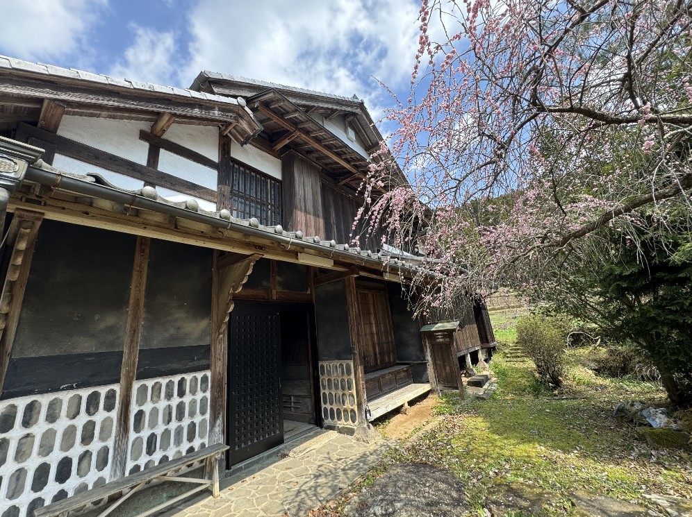 山あいの自然に囲まれた築古の日本家屋(古民家)の外観。土壁と木組みが残る歴史ある住まいが、長年の空き家として活用されずにいた様子が伺える。春の梅が咲く庭先で、空き家の現地調査を行い、古民家再生や空き家活用の可能性を検討するための参考事例。実家の空き家問題や売却・再生に悩む人向けの専門的な空き家資産の見直しを伝える文脈に適した写真。