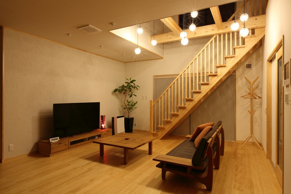吹き抜けのあるLDK空間に、木製階段とペンダント照明を組み合わせた住宅内観。高天井の吹き抜けに多灯配置した照明が上下階をやさしくつなぎ、無垢材の床と家具が心地よい明るさと落ち着きを生むリビング空間。