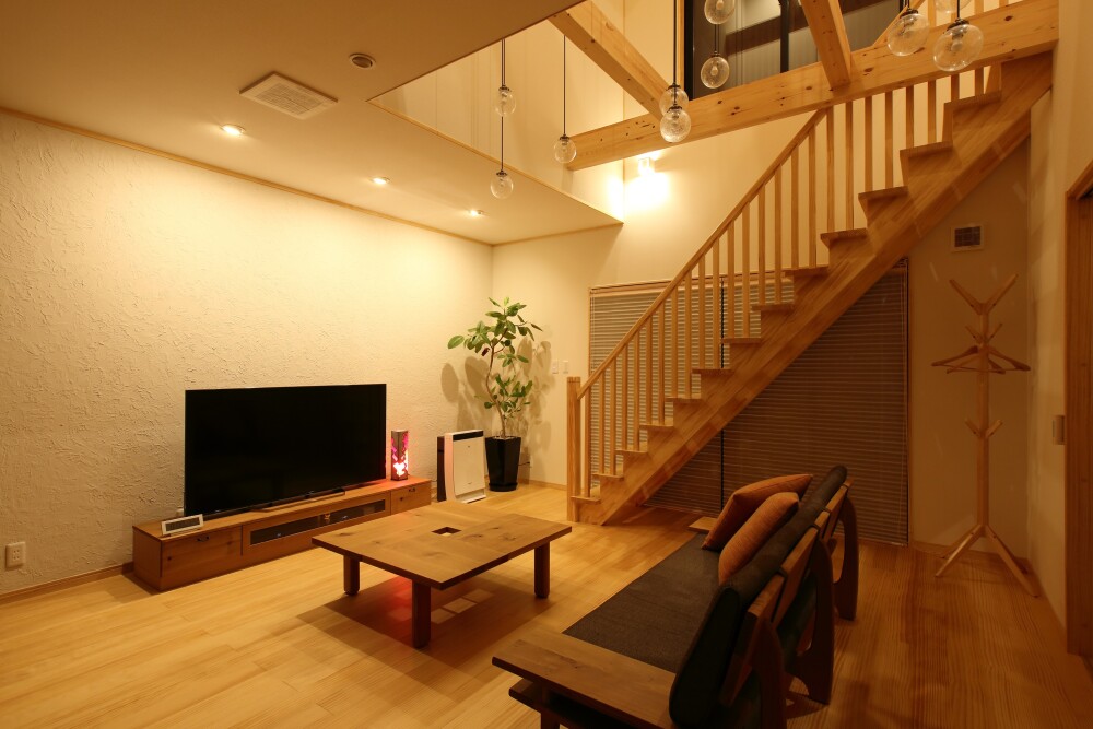 吹き抜けのあるLDK空間に、木製階段とペンダント照明を組み合わせた住宅内観。高天井の吹き抜けに多灯配置した照明が上下階をやさしくつなぎ、無垢材の床と家具が心地よい明るさと落ち着きを生むリビング空間。