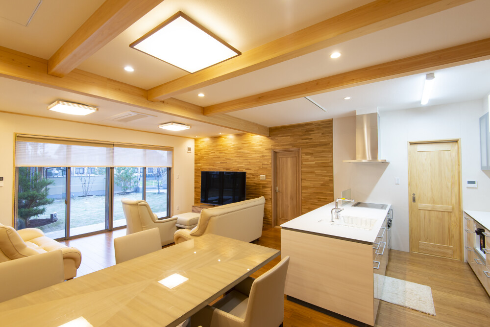 和モダン住宅のLDKをダイニング・キッチン側から見た空間構成。無垢材の梁と木質感のある天井、自然素材を活かしたインテリアにより、リビング・ダイニング・キッチンが一体となった設計。庭へと視線が抜ける開口部と回遊性のある間取りが、建築家による上質で落ち着いた和モダンの暮らしを演出している。