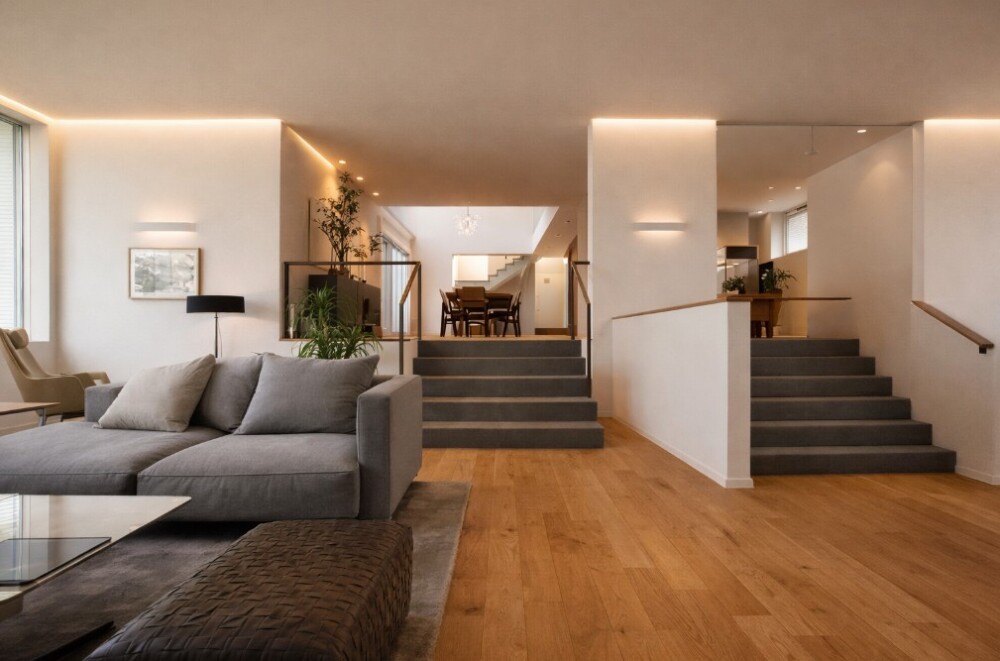 間取りに正解を求めるのではなく暮らしの最適解を考える設計思想を体現した、ホテルライクでモダンな注文住宅のLDK空間。グレーのソファとオークのフローリング、連続するコーニス間接照明、壁手摺の吹抜け階段が、家族の動線と自然なつながりを丁寧にデザインしている。やまぐち建築設計室が提案する、家族関係と時間の流れを大切にした住まいの実例。