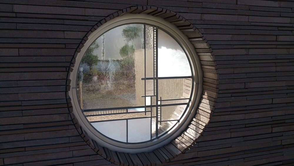 外壁に設えた円形窓にステンドグラスを組み込んだ和モダン住宅の外観ディテール。石調外壁と円窓の陰影に、幾何学模様の色ガラス越しの自然光が重なり、品格と奥行きを感じさせる建築デザイン。