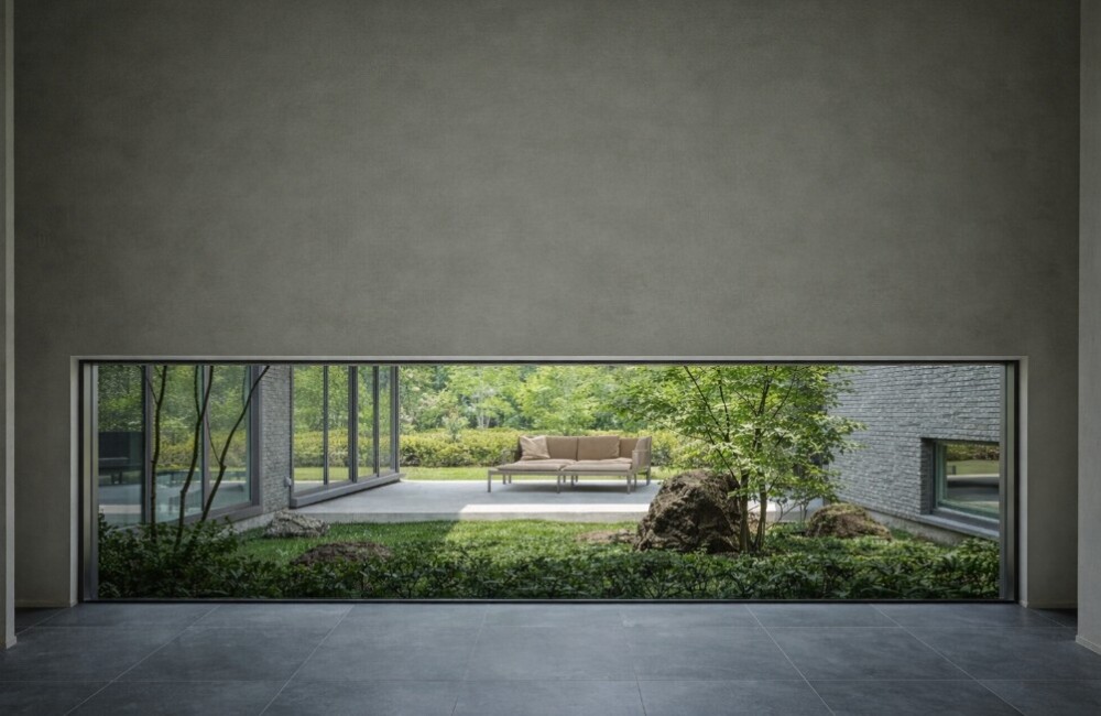 地窓越しに中庭を望む和モダン住宅の空間。グレーのタイル床と壁に囲まれた静かな室内から、植栽と石を配した中庭とテラスが緩やかにつながり、心を落ち着かせる余白のある暮らしを表現した建築設計。