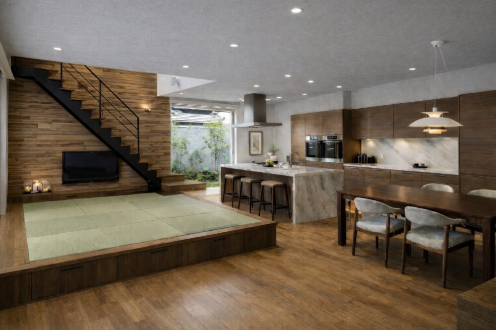 和モダンとホテルライクな美意識を融合させたLDK空間。 一般のLDK床より約350mm高く設けた畳の小上がりは、緑の畳を採用し、床座でくつろぐ日本的な居場所を現代的に再解釈している。 天井は薄いグレーのクロス仕上げとし、木質建材とのコントラストで空間に奥行きと落ち着きを与える構成。 テレビ背面と階段奥の壁には、建材メーカーWOODONEのDESIGN WALL「粋」を用い、素材感と陰影が際立つ上質なフォーカルポイントを形成。 キッチン・ダイニング・小上がりが緩やかにつながることで、家族の距離感と視線の抜けを丁寧に設計した、建築家による和モダン住宅の実例。 暮らしの質と時間の過ごし方を重視した、奈良県の設計事務所 やまぐち建築設計室 による住まい提案。