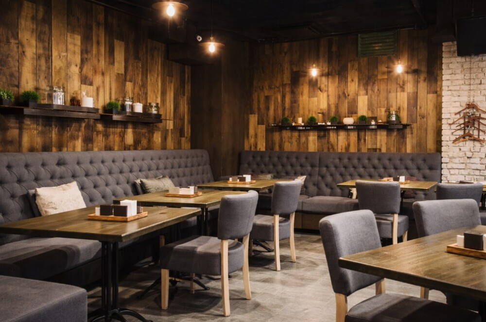 奈良県で個人経営・脱サラ開業を想定したカフェ空間事例。木目の壁とダークトーンのインテリア、グレーのソファ席と木製テーブルを組み合わせた落ち着いた店内デザイン。小規模カフェでも回りやすい客席配置と厨房動線を意識した設計例で、長く続くカフェ経営を支える空間づくりを表現している。