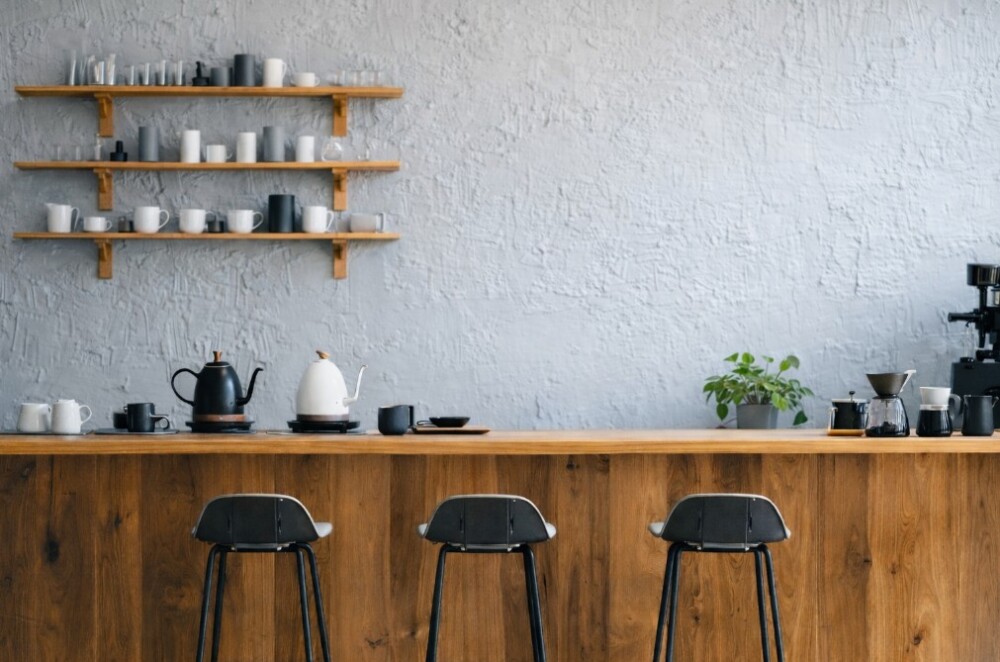 奈良県でカフェ開業・店舗設計を検討する個人オーナー向けの空間イメージ。木目仕上げのカウンターとスチール製ハイチェアを配置したコーヒー専門店の内観デザイン。壁面に設けた無垢材の棚にはドリップポット、サーバー、カップを整理収納し、エスプレッソマシンを含めた効率的な厨房動線を確保。カウンター端に配置した観葉植物ポトスが柔らかな印象を与え、長く続く個人経営店を想定したミニマルで機能的な店舗設計の事例イメージ。