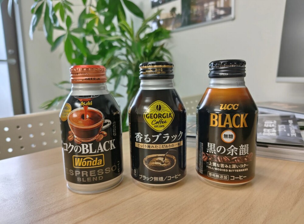 木目のテーブルの上に並ぶ3本のブラック無糖缶コーヒー。左からアサヒ「WONDA モーニングショットBLACK」、ジョージア「香るブラック」、UCC「BLACK 黒の余韻」。いずれも黒を基調としたパッケージだが、ロゴの書体や色彩設計、コピー表現の違いによって「力強さ」「香り」「余韻」といった異なる味覚イメージを想起させるデザイン比較写真。背景には観葉植物と自然光が差し込む室内空間が広がり、パッケージデザインが与える印象の違いと、ブランド設計の重要性を示している。奈良で飲食店開業や店舗デザインを検討する際に、空間設計やブランディングが体験価値を左右することを象徴する一枚。
