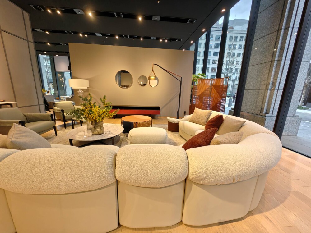 ポルトローナ・フラウ大阪ショールームのラウンジ空間。曲線的なフォルムのベージュソファを中心に、丸テーブルとフロアランプ、アクセントカラーのクッションを配置し、自然光と間接照明が調和するホテルライクで上質なリビングインテリア。家具と空間設計が一体となった居心地の良い住まいの提案。