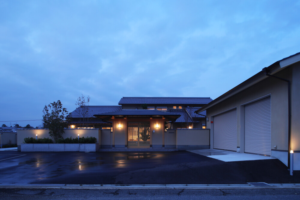 和モダン住宅の外観と門構えの夜景。奈良の注文住宅事例として、数寄屋の意匠を取り入れた玄関アプローチと塀、柔らかな間接照明が迎える落ち着いた住まい。ビルトインガレージと外構計画が一体となり、上質な暮らしを演出する建築家設計の住宅。