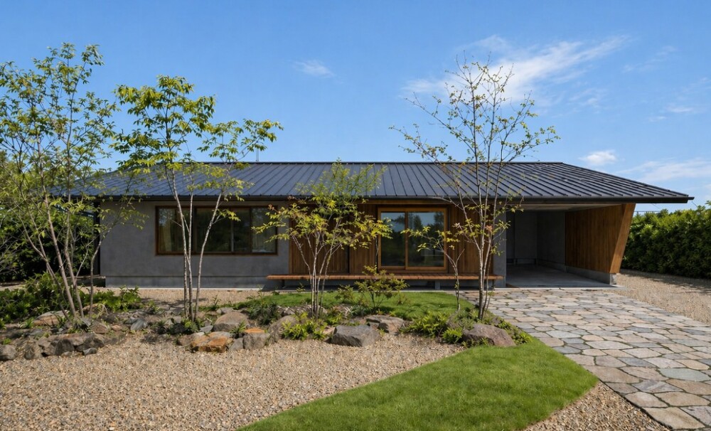 奈良の気候風土に寄り添って設計された和モダンの平屋住宅外観。濃いグレーのジョリパット仕上げの外壁とガルバリウム鋼板の屋根が調和し、深い軒が日差しをやわらかく遮る。木の質感と石張りアプローチ、植栽が一体となり、静けさと上質な暮らしを感じさせる外構デザイン。