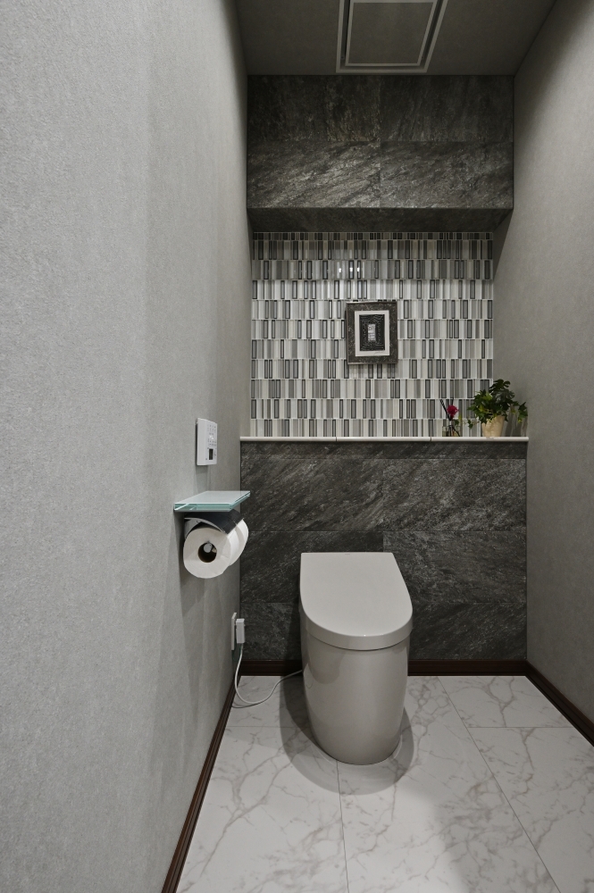 ホテルライクな高級トイレ空間の設計事例。グレートーンの左官壁と石目調タイル、モザイクタイルのアクセントウォールが陰影と奥行きを生み出し、落ち着きのあるラグジュアリーな雰囲気を演出。フロート感のある床仕上げとシンプルな設備配置により、清潔感と静けさを両立。和モダン住宅における上質な水まわり空間デザインの実例。