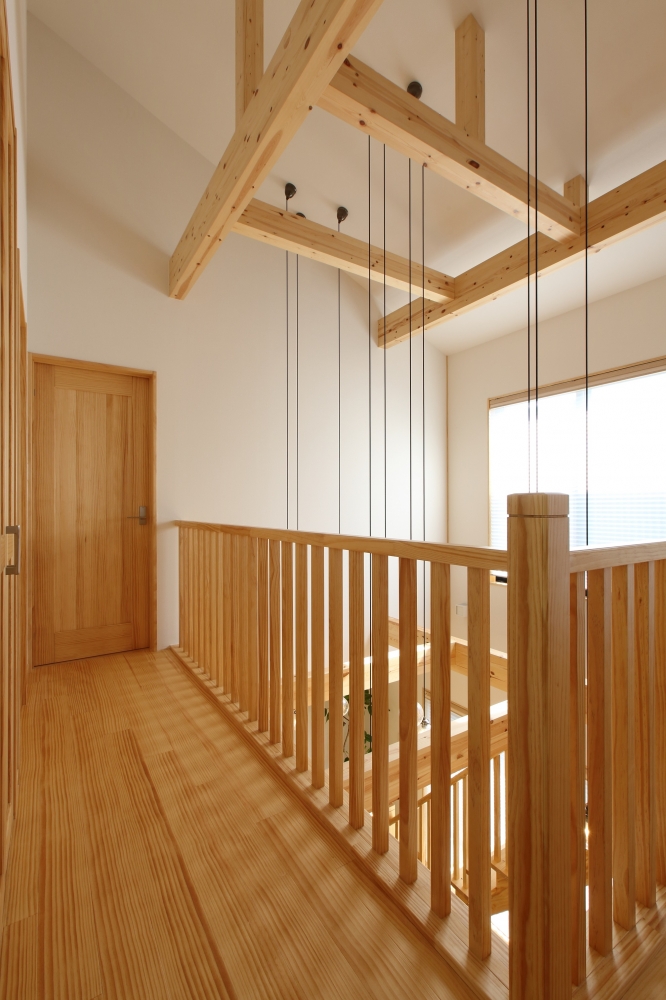 和モダン住宅の2階ホールと吹き抜け空間。無垢材の手すりと床、現しの梁が連続し、上階から下階へと視線と光が通り抜ける設計。ペンダント照明のコードが縦のラインを強調し、空間にリズムと奥行きを与える。奈良の注文住宅における、上下階の一体感と静かな居心地を実現した上質な住まいの施工事例。