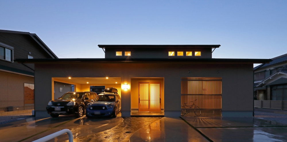 夕景に浮かび上がる、ビルトインガレージを備えた和モダン住宅の外観デザイン。深い軒と水平ラインを強調した落ち着きある佇まいに、木格子から漏れるあたたかな光が上質な住まいの気配を演出。ブラックとグレージュを基調にした外壁構成と、光の陰影を丁寧に計画した外観は、和の趣とホテルライクな美意識を融合させた設計事例。雨の日でも快適なハーフビルトインガレージや、プライバシーに配慮した開口計画により、暮らしやすさと美しさを両立。和モダンの家、高級注文住宅、ガレージハウス、デザイン住宅、上質な暮らしを求める家づくり検討者に向けた住まい提案。