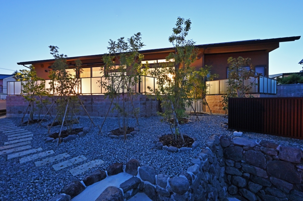 奈良県の和モダン注文住宅の夕景外観。石と砂利の庭に植栽を配置し、建物の大開口から漏れる室内の灯りが外構と調和。深い軒と水平ラインが美しい平屋風の住まいで、内外がつながる設計により静けさと上質な暮らしを実現した住宅事例。