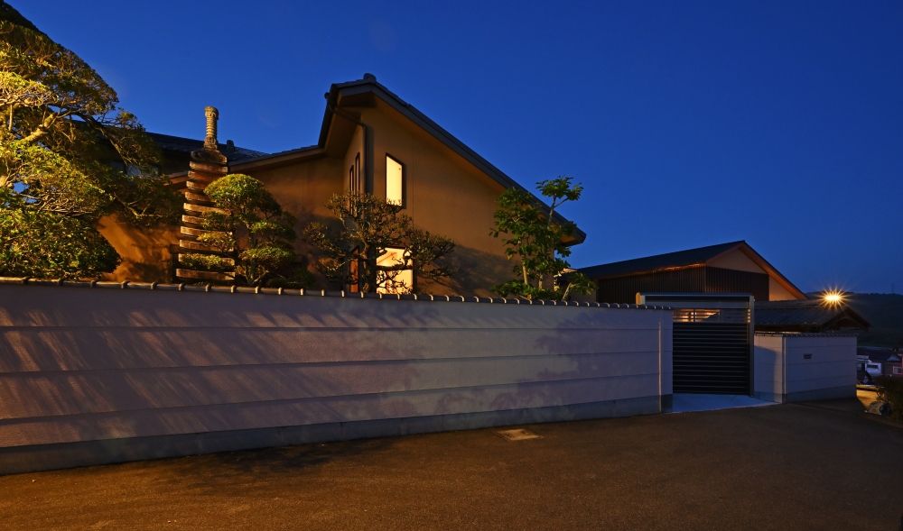奈良県の和モダン注文住宅の夜景外観。塀に囲まれた庭と石塔、手入れされた植栽がライトアップされ、建物のやわらかな室内灯と調和。伝統的な屋根形状と落ち着いた外壁が特徴で、外構と建築が一体となり静けさと品格ある暮らしを実現した住宅事例。