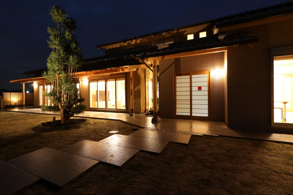奈良県のやまぐち建築設計室が設計した数寄屋造りの高級和風住宅外観（夜景）。瓦屋根と深い軒に守られた玄関と格子戸が印象的で、室内からの暖かな光が庭と石張りのアプローチに広がる設計。植栽と外構照明が陰影を生み、内外の一体感と静けさを演出する建築家設計の和モダン注文住宅事例。