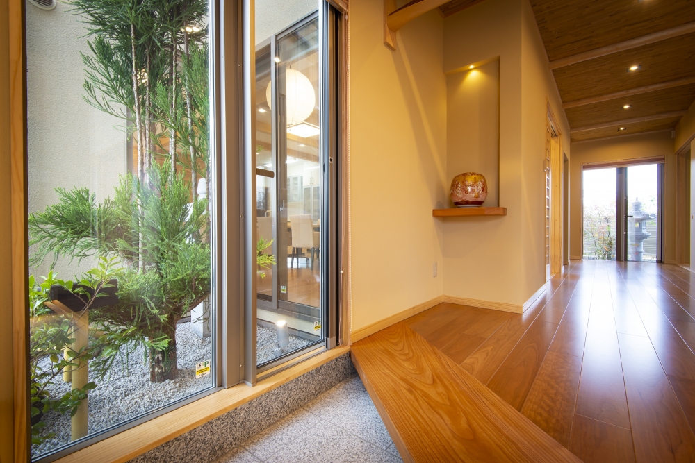 奈良県のやまぐち建築設計室が設計した数寄屋風和モダン住宅の玄関ホールと坪庭の内外一体空間。大開口の窓越しに植栽を取り込み、無垢フローリングと式台、木天井が温かみのある空間を構成。奥へと続く廊下と庭の景色が視線の抜けを生み、光と陰影が調和する高級注文住宅の設計事例。和風住宅・和モダン住宅の玄関デザイン。