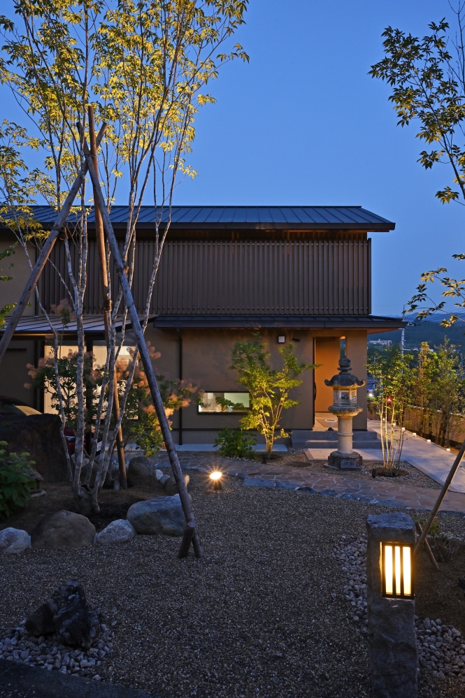 奈良県の和モダン注文住宅の夜景外観。ライトアップされた庭と石灯籠、植栽が建物と調和し、木格子と深い軒が陰影を際立たせる住まい。外構と建築が一体となり、光と影の演出によって上質で静かな暮らしを実現した住宅事例。