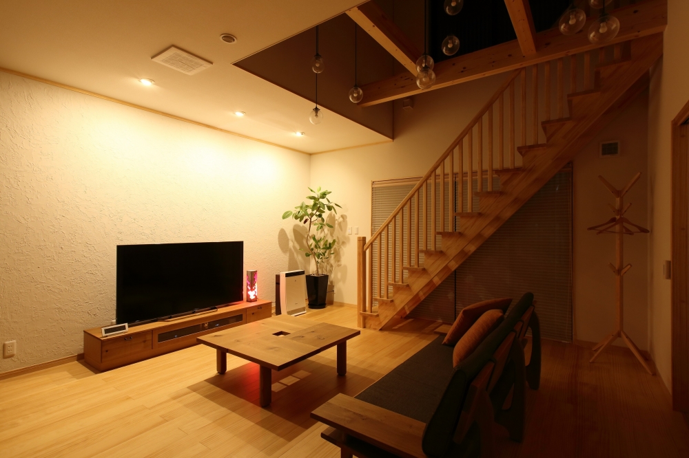 奈良の注文住宅における和モダンのリビング空間。吹き抜けとリビング階段を中心に、無垢材の床・梁・手すりを活かした設計に、ペンダント照明と間接照明を組み合わせた照明計画を採用。夜の時間帯でも温かみのある光が広がり、ホテルライクで上質なくつろぎを実現する高級住宅の内観デザイン事例。