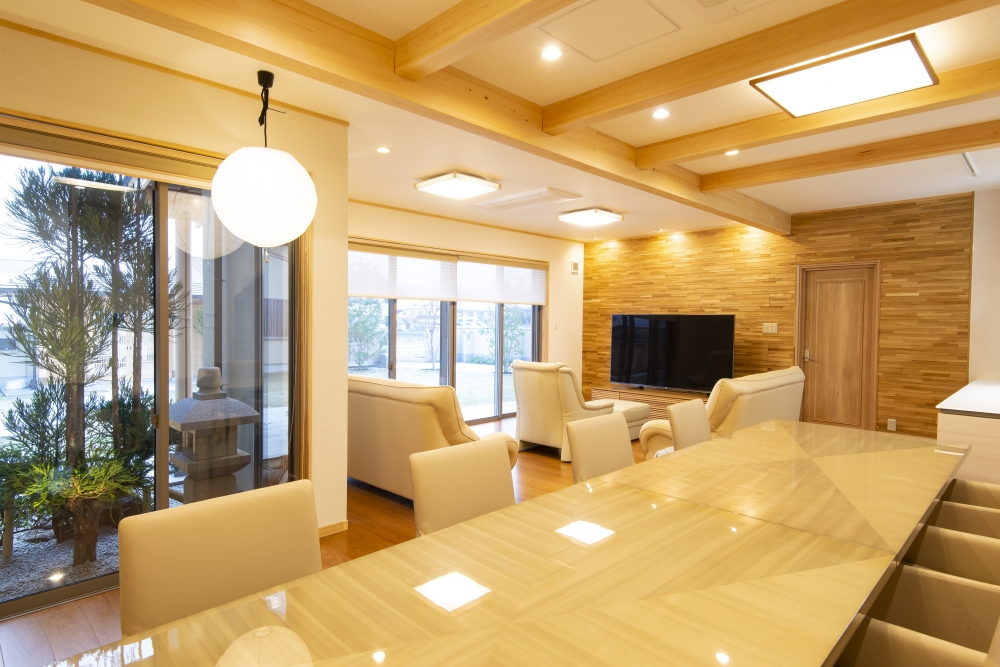 奈良県のやまぐち建築設計室が設計した和モダン住宅のLDK空間。無垢材の梁と木質アクセントウォールを活かしたリビングに、長尺ダイニングテーブルと対面キッチンを配置。大開口サッシから庭と中庭を望む設計で、採光・通風・視線の抜けを確保。自然素材の温もりとホテルライクな上質感を融合した高級注文住宅の実例。
