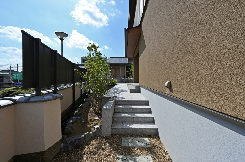 奈良県の和モダン住宅の外構アプローチ。塀と建物に挟まれた動線に、石張りの階段と植栽を配置し、外から内へと連続する空間を構成。深い軒と落ち着いた外壁が陰影を生み、視線をコントロールしながら静けさと安心感を高める注文住宅の設計事例。