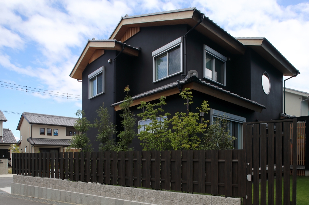 黒を基調とした和モダン住宅の外観と、角地の特性を活かした外構・植栽計画。深い軒と丸窓、日本建築らしい水平ラインが落ち着いた佇まいを生み出し、木調フェンスと庭木によって外からの視線をやわらかく整えている。どの方向から見ても美しく見えるよう建築と庭を一体で設計し、街並みに調和しながらも静かな存在感を放つ住まい。和風住宅・純和風の家・和モダン住宅・黒い外観の家・ホテルライクな家・デザイナーズ住宅を求める家づくり検討者へ向けた、上質な注文住宅事例。光・陰影・植栽・外構まで丁寧に整えた和モダンの住空間。