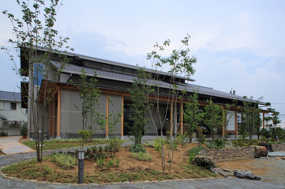奈良県の建築家設計による高級和モダン住宅の外観と庭の一体デザイン。 深い軒と木格子、瓦屋根の水平ラインに、石積みと植栽を組み合わせた外構が調和し、四季を感じる庭と暮らす注文住宅。