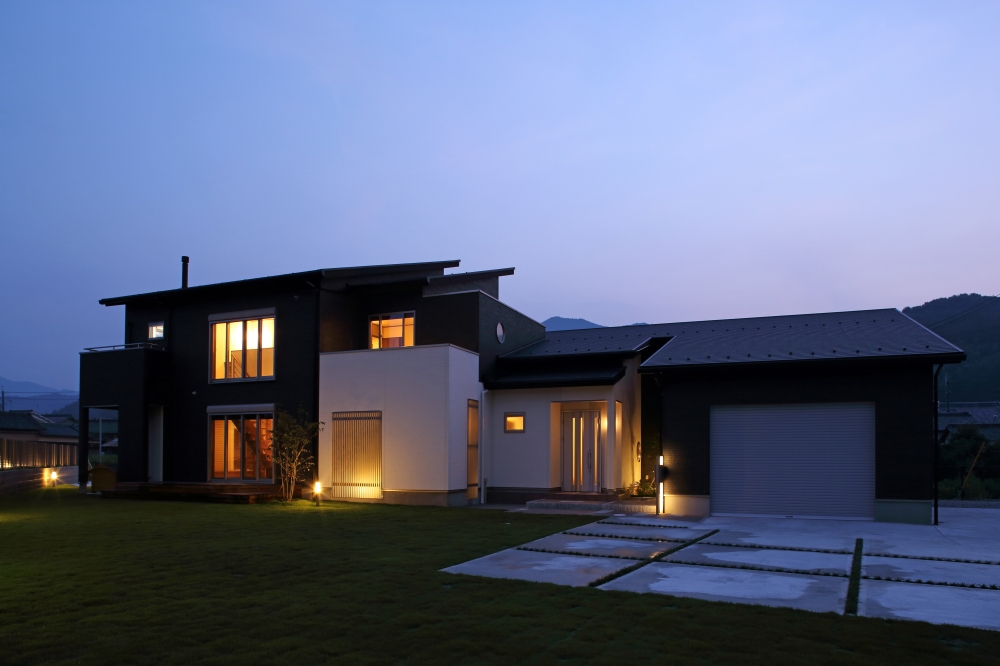 夕景に佇む和モダン住宅の外観。ブラックとホワイトのコントラストに、室内から漏れる柔らかな光が上質なホテルライクな雰囲気を演出。ビルトインガレージと庭が一体となり、機能性と美しさを両立した注文住宅の設計事例。