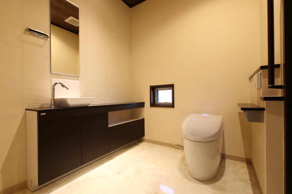 奈良の注文住宅における二階トイレ空間。手洗いカウンターと一体化した広めのトイレに、落ち着いた木質キャビネットと間接照明を組み合わせ、ホテルライクで上質な空間を実現。小窓からの採光と清掃性に優れた床材により、快適性と機能性を兼ね備えた和モダン住宅の設計事例。