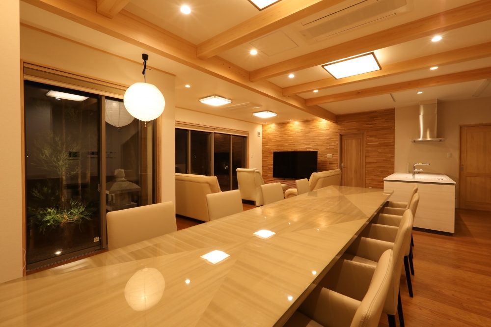 奈良県のやまぐち建築設計室が設計した数寄屋風の高級和モダン住宅のLDK空間。無垢材の梁を現しにした天井構成と、間接照明・ダウンライトによる柔らかな光環境が特徴。大開口サッシ越しに中庭とつながるダイニングキッチンは、アイランドキッチンと一体化し、家族の動線と視線の広がりを両立。高級和風住宅における上質な団欒空間の設計事例。