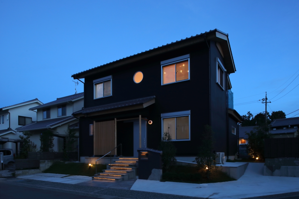 　夕暮れの街並みに静かに佇む、黒を基調とした和モダン住宅の夜景外観。角地の特性を活かした建築計画により、どの方向から見ても整った美しいファサードを形成。深い軒と木格子、丸窓、日本建築らしい水平ラインが、やわらかな灯りと共に陰影豊かな表情を生み出している。階段アプローチの間接照明や外構照明が、ホテルライクな上質感と安心感を演出し、昼とは異なる落ち着きある景観を実現。和風住宅・純和風の家・和モダン住宅・黒い外観の家・デザイナーズ住宅・高級感のある注文住宅を求める家づくり検討者へ向けた住まい事例。