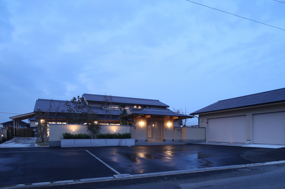 奈良県のやまぐち建築設計室が設計した数寄屋造りの高級和風住宅外観。瓦屋根と深い軒、格子戸のある門構えが美しい迎えの空間を形成し、塀内に広がる植栽と間接照明が上質な陰影を演出。広々とした駐車スペースと一体となった外構計画により、格式と機能性を両立した建築家設計の注文住宅事例。
