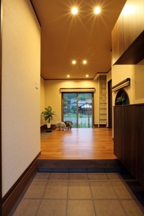 和モダン住宅の玄関ホール。木質フローリングと落ち着いた色合いの内装に、ダウンライトのやわらかな照明が陰影を生み出す設計。大開口サッシから外の景色と自然光を取り込み、視線の抜けと奥行きを感じる、ホテルライクで上質な玄関空間。