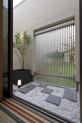 畳の和室から望むモダンな中庭空間。白砂利と飛び石、植栽を配した外構に縦格子を設けることで、外部からの視線をやわらかく遮りながら光と風を取り込む設計。障子越しに感じる自然の気配と、室内外が緩やかにつながる構成により、和モダン住宅やホテルライクな暮らしを求める方に適した上質な住まいの一例。