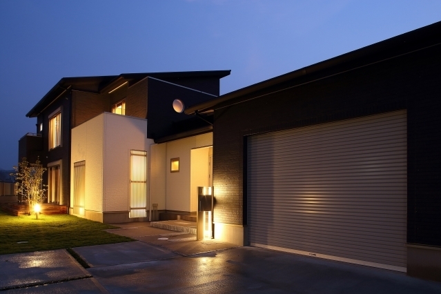 夜景に浮かび上がるビルトインガレージ付き和モダン住宅の外観。黒と白のコントラストが美しいファサードに、間接照明と外構照明が温かみのある光を演出。玄関アプローチからガレージまで一体的に計画された動線設計により、機能性とデザイン性を両立。芝生の庭と建物が調和し、ホテルライクで上質な暮らしを実現する注文住宅。