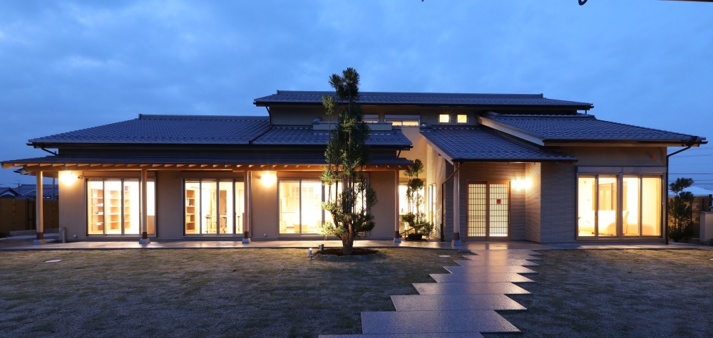 奈良県のやまぐち建築設計室が設計した数寄屋造りの高級和風住宅外観（夕景）。瓦屋根と深い軒が連続する水平ラインが美しい平屋建て住宅で、大開口の掃き出し窓から室内の暖かな灯りが庭へと広がる設計。石張りのアプローチと庭の植栽、外構照明が一体となり、内と外を緩やかにつなぐ上質な和モダン住宅の建築事例。