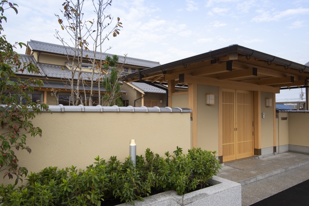 奈良県のやまぐち建築設計室が設計した数寄屋風和モダン住宅の門構え外観（斜め視点）。木製格子の門扉と深い軒、土塀と植栽が調和した落ち着きある外構デザイン。瓦屋根と木の質感が上質な陰影を生み、内側の庭と建築へとつながる奥行きある空間構成が特徴の高級注文住宅事例。和風住宅・和モダン住宅における品格あるエントランス設計。