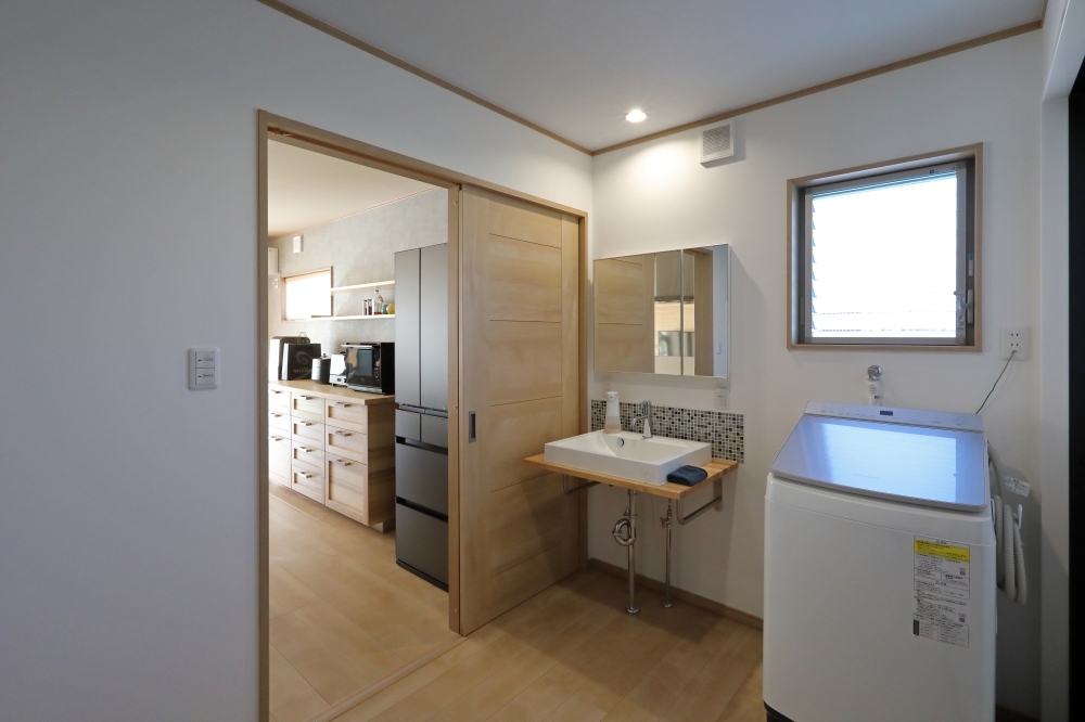 キッチンと玄関ホールをスムーズにつなぐ、二方向動線を採用した和モダン住宅の洗面脱衣室。家事動線と帰宅動線を効率的に整理し、洗濯・身支度・収納までを一体的に整えた暮らしやすい空間設計。木の温もりを感じる建具とシンプルな洗面台、モザイクタイルのアクセントが、ホテルライクで清潔感のある雰囲気を演出している。キッチン側からも廊下・玄関ホール側からもアクセスできる回遊動線により、家族の動きが重なりにくく、毎日の家事負担を軽減。和モダンの家・洗面脱衣室・回遊動線・家事ラク動線・高級注文住宅・暮らしやすい間取りを求める方に向けた住宅設計事例。