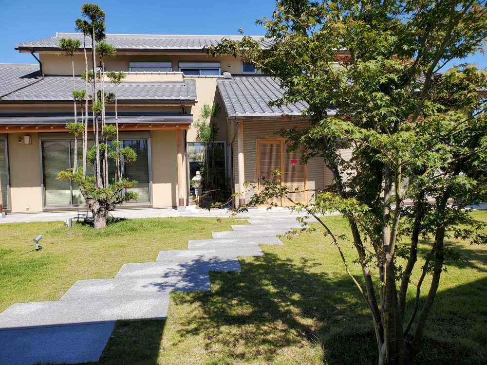 奈良県のやまぐち建築設計室が設計した数寄屋風和モダン住宅の玄関アプローチと庭の外観。芝生の庭に石畳の動線を設け、植栽と調和した落ち着きある景観が特徴。深い軒と瓦屋根の陰影、木製格子の玄関が上質な迎えの空間を演出。内外のつながりと時間の流れを大切にした高級注文住宅の設計事例。