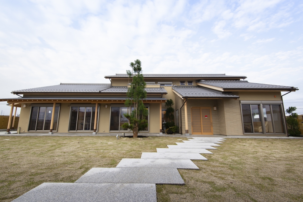 奈良県のやまぐち建築設計室が設計した数寄屋風和モダン住宅の庭側全景外観。芝生の広がる庭に石貼りのアプローチを設け、玄関へと導く動線計画が特徴。瓦屋根と深い軒、木製格子の玄関、連続する開口部が内外のつながりを生み出し、庭と一体となった開放的で上質な住環境を実現した高級注文住宅の設計事例。和風住宅・和モダン住宅の庭一体型デザイン。