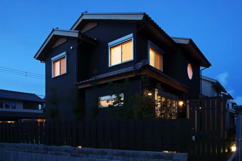 夜の街並みに静かに佇む、黒を基調とした和モダン住宅の夜景外観。深い軒と木の温もりを感じる軒天、丸窓、日本建築らしい水平ラインが、柔らかな室内灯によって美しく浮かび上がる。木調フェンスと植栽、外構照明を一体で整えることで、プライバシーを守りながらも開放感と上質感を演出。ホテルライクな雰囲気と和風住宅の静けさを融合したデザインで、純和風の家・黒い外観の家・和モダン住宅・デザイナーズ住宅を求める家づくり検討者へ向けた住まい事例。建築・照明・外構計画を丁寧に調和させた夜景の美しい注文住宅。