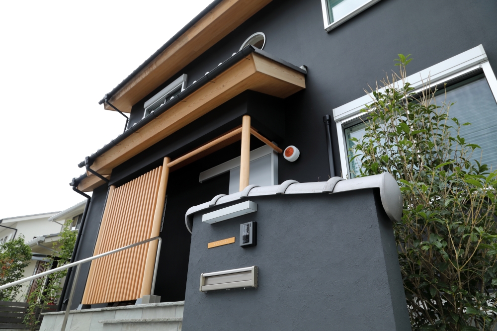 黒を基調とした和モダン住宅の正面外観。深い軒と無垢材の軒天、縦格子、丸窓が日本建築らしい趣を生み出し、現代的なデザインと伝統美を融合した住まい。門塀には瓦をあしらい、外構と建築を一体で整えることで、街並みに落ち着きと品格を与えている。ホテルライクな上質感と、和風住宅ならではの静けさを感じるデザインで、純和風の家・和モダン住宅・黒い外観の家・デザイナーズ住宅・高級感のある注文住宅を求める家づくり検討者に向けた建築事例。光と陰影、素材感、外観バランスまで丁寧に設計された住まい。