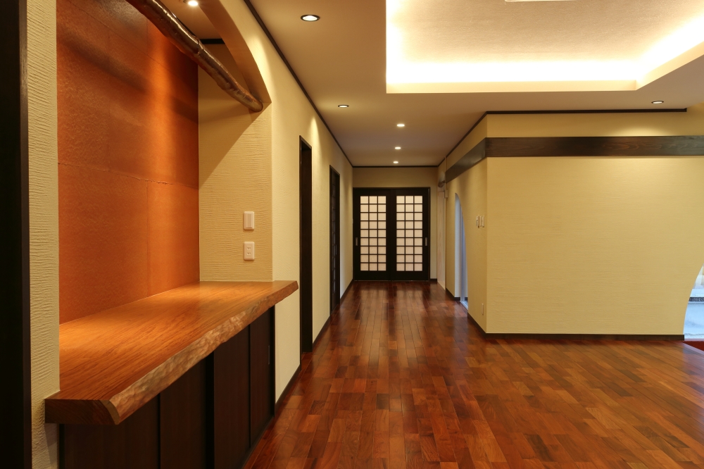 奈良の注文住宅における玄関ホールから中庭回廊へとつながる空間設計。無垢フローリングと木質カウンターを配した上質な内装に、間接照明とダウンライトで陰影を整え、奥行きのある動線を演出。中庭を囲う回廊へと視線が抜けることで、内外一体の広がりを感じる和モダン住宅の設計事例。