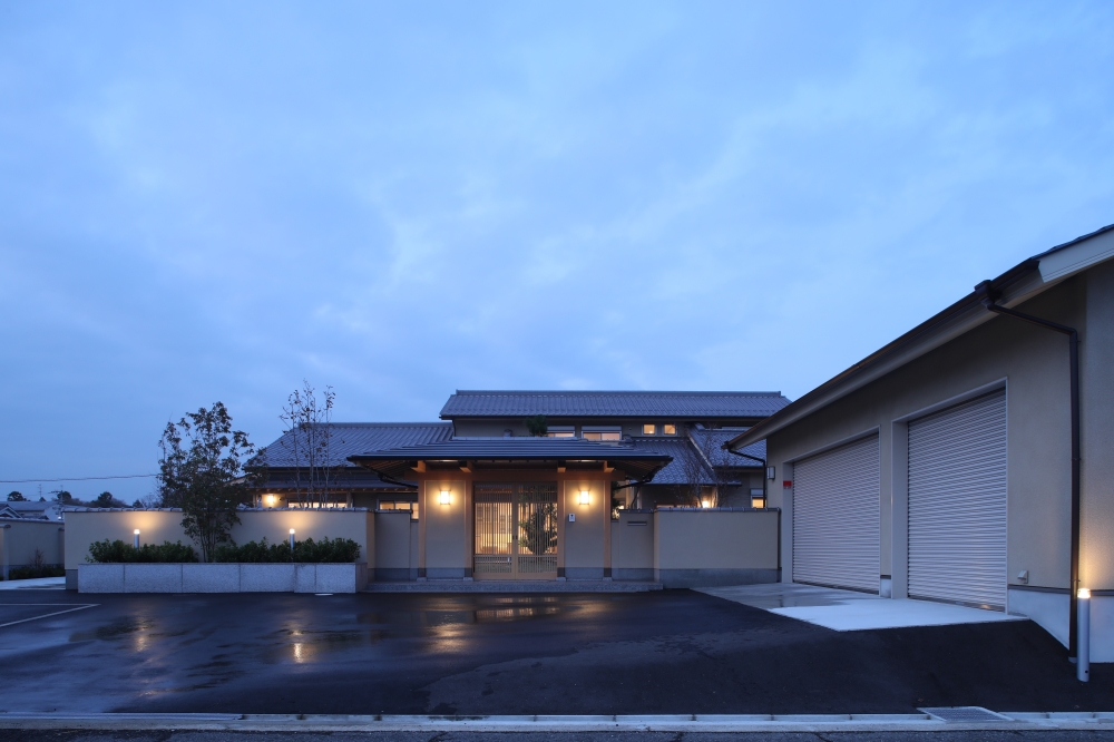 奈良県のやまぐち建築設計室が設計した数寄屋造りの高級和風住宅外観（夕景）。瓦屋根と深い軒を持つ母屋と、格子戸のある門構えを塀で囲った外構計画が特徴。間接照明と植栽が織りなす陰影が、落ち着きと品格を演出し、広い駐車スペースとガレージを備えた機能性の高い和モダン注文住宅の建築事例。