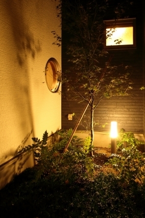 夜の中庭に設けた和モダン住宅の外部空間。植栽と外壁にあたる間接照明がやわらかな陰影を生み、丸窓や小窓から漏れる光と調和して静けさと奥行きを演出。足元のライティングと自然素材が心を落ち着かせ、ホテルライクな上質感と癒しを感じる設計。外と内のつながりを意識し、時間の質と感性を豊かにする中庭空間。
